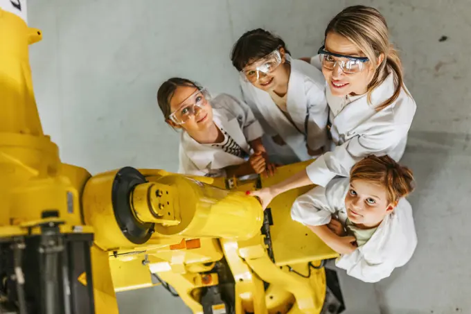 Top view of children learning robotics in Elementary school, standing with female teacher by robotic arm. After-school robotics club, or field trip to real robotics laboratory. Portrait of children learning robotics in Elementary school, standing with female teacher. Young students building robot in after school robotics club. Field trip to real robotics laboratory. model released