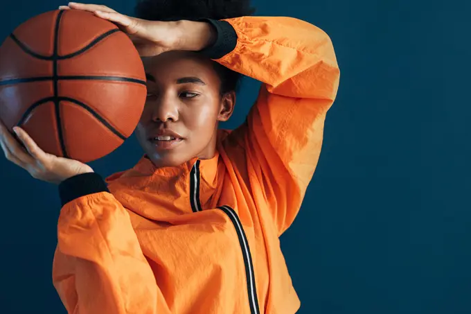 Close up studio shot of young female basketball player in orange sportswear posing with ball. Portrait of a professional basketball player over blue background. Close up studio shot of young female basketball player in orange sportswear posing with ball. Portrait of a professional basketball player over blue background. Copyright: xxArtemxVarnitsinx